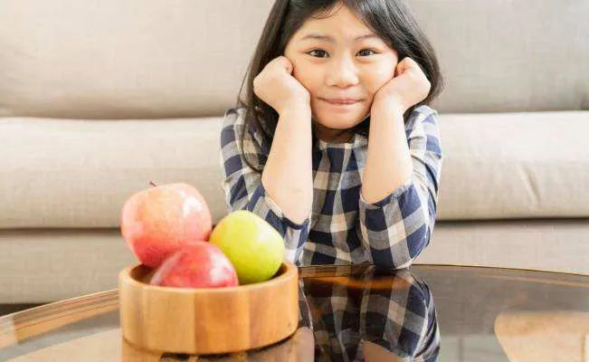 A young girl sits at a glass coffee table with a wooden bowl of red and green apples, hands on her cheeks.