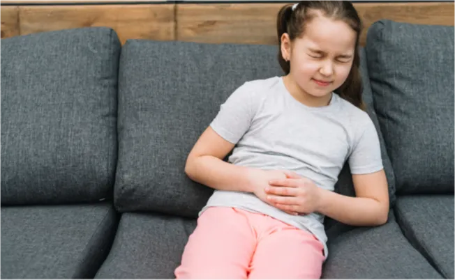 A young girl sits on a gray couch with her hands clasped over her stomach, appearing uncomfortable.