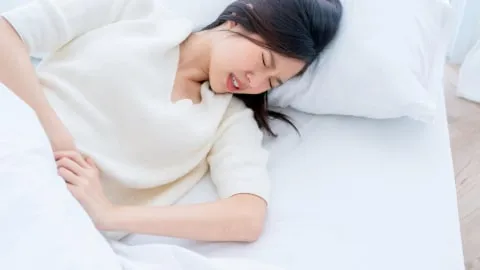 A woman lying in bed in a brightly lit room, appearing to be in pain or distress, with her eyes closed and hand grasping the bed sheet.