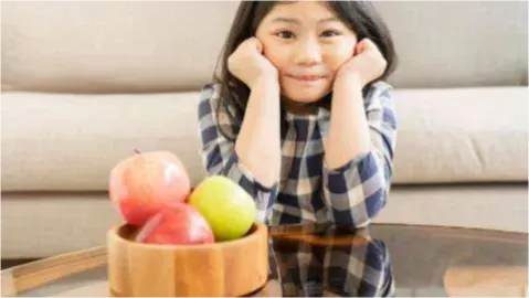 A young girl sits at a glass coffee table with a wooden bowl of red and green apples, hands on her cheeks.