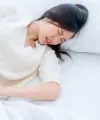 A woman lying in bed in a brightly lit room, appearing to be in pain or distress, with her eyes closed and hand grasping the bed sheet.