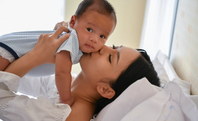 Mother kissing baby as she carries them while lying on bed.